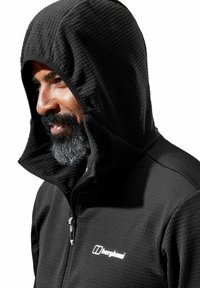 Zwarte zip-up jack met een gestructureerde capuchon, met een soepele stoflichaam en contrasterende stiksels. Logo zichtbaar op de borst.