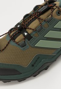 Bėgimo bateliai takams rudos ir žalios spalvos, su tekstūruotu tinkleliu viršuje, GORE-TEX prekės ženklu, juodais ir oranžiniais raišteliais bei apsauginėmis detalėmis.