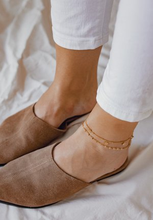 Beżowe zamszowe buty typu slip-on ze kwadratowym noskiem. Białe rurki jeansowe. Złote bransoletki na kostkę z małymi dekoracyjnymi charmsami. Widoczna gładka skóra.