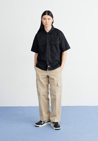 Sort kortærmet skjorte med knapper og to brystlommer, parret med tan cargo-bukser. Sorte sneakers med hvide detaljer, stående på blåt gulv.