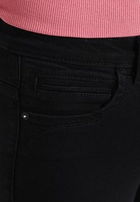 Jeans de mezclilla negra con un bolsillo frontal que presenta un sutil detalle de costura, combinados con una cinturilla de color rosa acanalada visible por encima de los jeans.