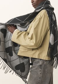 Gray and black patterned scarf featuring a heart design, with fringed edges. Worn over a beige jacket, showcasing its soft texture.