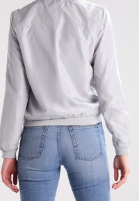 Blouson aviateur gris clair avec une texture lisse, des poignets élastiques et un ourlet côtelé. Porté sur un jean bleu ajusté, montrant des coutures détaillées.