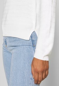 Close-up of a person wearing a white textured sweater and light blue denim jeans, with the hand relaxed by the side.