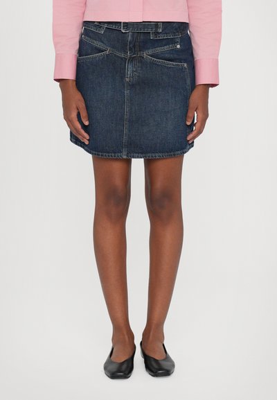 Denim nederdel i mørkeblå, med en tætsiddende talje, frontlommer og en let buet kant. Sammen med enkle sorte flade sko.
