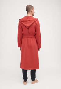 Rød teksturert badekåpe med hette, belted i midjen, lange ermer og ned til midten av leggen, matchende med mørke bukser og bare føtter.