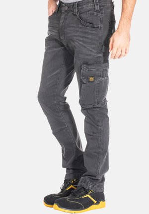 Pantalons cargo gris avec une coupe slim, cinq poches et une étiquette avec logo jaune. Portés avec des chaussures de sport noires et jaunes.