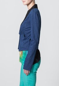 Blå denimjacka i stil med svart krage, med en figursydd design och långa ärmar, lagd över en skjorta med tie-dye-mönster och gröna byxor.