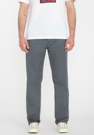 Muž oblečený v sivých nohaviciach s rovnými nohavicami, bielych teniskách a bielom tričku s logom Volcom, stojaci pred jednofarebným bielym pozadím.