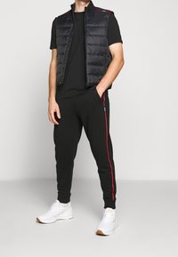 Sort polstret vest med røde detaljer, parret med en sort t-shirt og sorte joggingbukser med røde sidestriber, samt hvide sneakers.