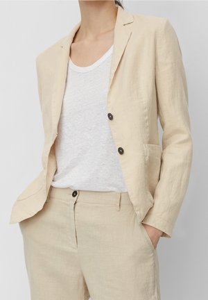Persona que lleva una chaqueta de lino beige y pantalones a juego con las manos en los bolsillos, combinados con una camiseta blanca lisa.