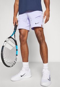 Vaaleanlilat tennishortsit, joissa on sivuventtiilit ja musta Nike-logo, yhdistettynä valkoisiin urheilukenkiin ja mustaan tennismailaan, jossa on siniset yksityiskohdat.