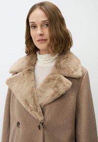Cappotto beige doppiopetto con collarino in pelliccia sintetica. Tessuto di lana testurizzato, grandi rever e bottoni scuri. Il modello indossa un dolcevita di colore chiaro.