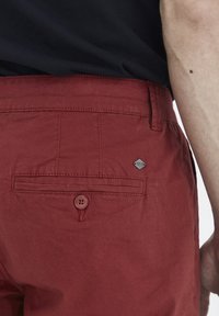 Pantalones rojos de algodón que presentan un bolsillo trasero con cierre de botón y un parche de logo en forma de diamante. La tela tiene una textura suave.