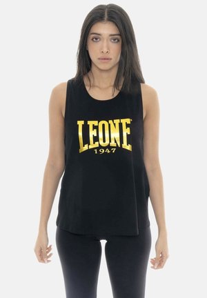 Jeune femme aux longs cheveux foncés portant un haut noir sans manches avec le texte doré « LEONE 1947 », debout devant un fond blanc uni.