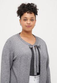 Harmaa neulecardigan, jossa on pyöreä pääntie, etuosassa siteen yksityiskohta ja kontrastisia mustia pystyraitoja sisäkerroksessa. Pehmeä materiaali, pitkät hihat.