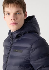 Giacca puffer blu navy con chiusura zip frontale, texture imbottita, cappuccio e un'etichetta nera sul petto. Materiale liscio con cuciture orizzontali.