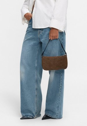 Persona che indossa jeans blu a gamba larga e una camicia bianca, mentre tiene una borsa in suede marrone, abbinata a scarpe nere con punta a stiletto.