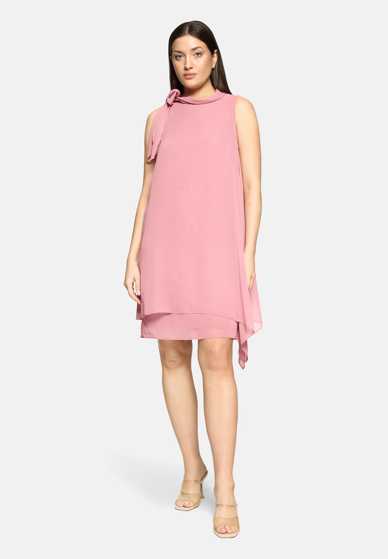 Robe rose sans manches avec un détail de nœud au niveau du décolleté, tissu fluide et ourlet asymétrique. Associée à des sandales transparentes.