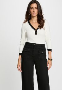 Top de manga larga blanco con ribetes negros y detalle de botones, combinado con pantalones de pierna ancha de talle alto negros con bolsillos delanteros.