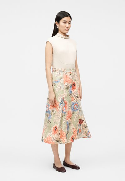 Γυναίκα με μακριά μαύρα μαλλιά, ντυμένη με αμάνικη μπεζ μπλούζα και φούστα midi με floral σχέδιο και ψηλή μέση, καθώς και σκούρα καφέ φλατ παπούτσια, στέκεται πλαγίως.