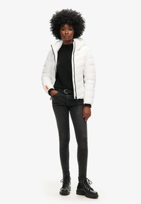 Doudoune blanche avec capuche et motif matelassé, pull noir, jeans skinny foncés et bottes noires à lacets ; présente des accents orange.