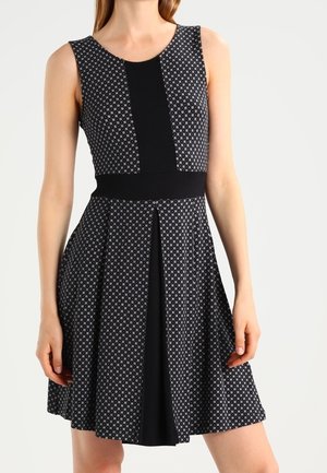 Czarna sukienka bez rękawów z małymi białymi geometrycznymi wzorami, z dopasowaną talią i centralną plisą z czarnego panelu, noszona przez stojącą osobę.