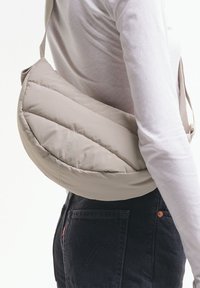 Henkilö, jolla on valkoinen pitkähihainen paita ja mustat farkut, kantaa beigeä, tikattua, puolikuun muotoista olkalaukkua.