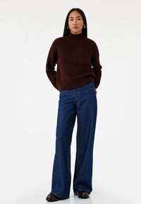 Brun strikket sweater med høj hals, parret med brede blå denimjeans. Sorte sko fuldender outfittet mod en hvid baggrund.