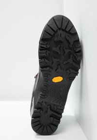 Mustaan vaelluskengän pohja, jossa on syvät, monisuuntaiset urat. Keskellä on keltainen Vibram-logo kestävyys- ja pidon takaamiseksi.