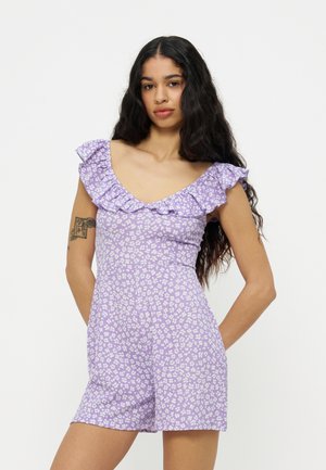 Žena s dlouhými tmavými vlasy, oblečená v lila květinovém overal s volánkovým odhaleným výstřihem, stojící se založenýma rukama za zády.