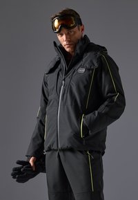 Chaqueta aislante negra con detalles de cremallera y snaps, presenta acentos en amarillo brillante, alto cuello y múltiples bolsillos con cremallera. Guantes sostenidos en la mano.