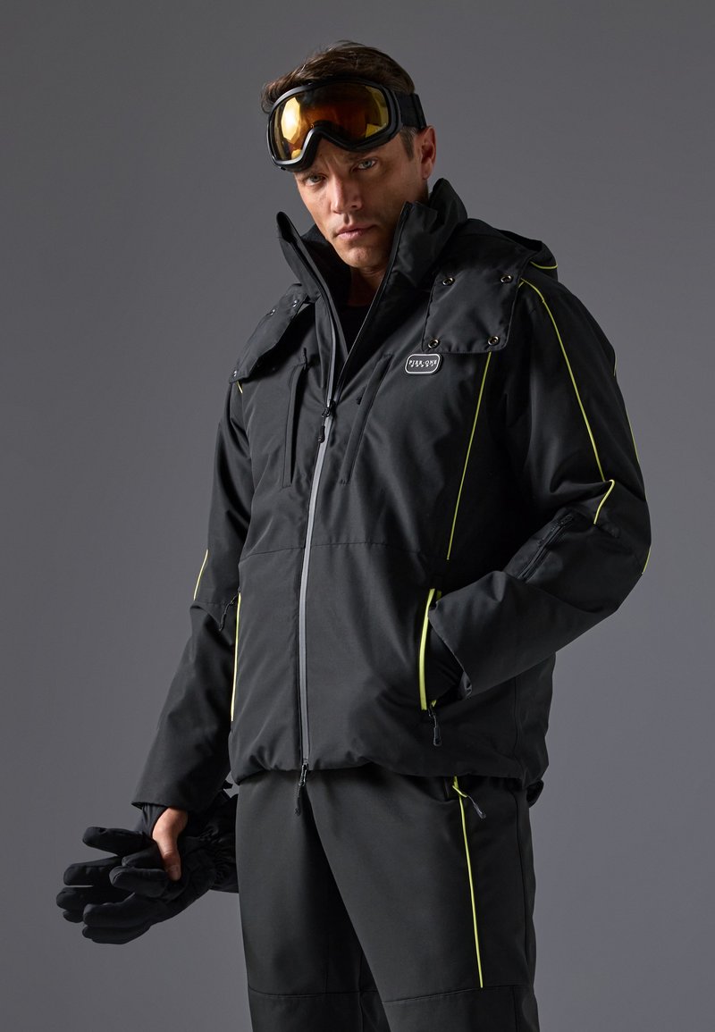 Chaqueta aislante negra con detalles de cremallera y snaps, presenta acentos en amarillo brillante, alto cuello y múltiples bolsillos con cremallera. Guantes sostenidos en la mano.