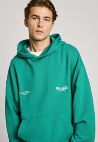 Sudadera con capucha verde hecha de algodón con una superficie texturizada. Presenta logotipos impresos en blanco en el frente y un ajuste relajado.
