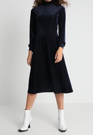 Woman wearing a long-sleeve black midi dress with a high neck paired with white ankle boots, standing against a plain light background.