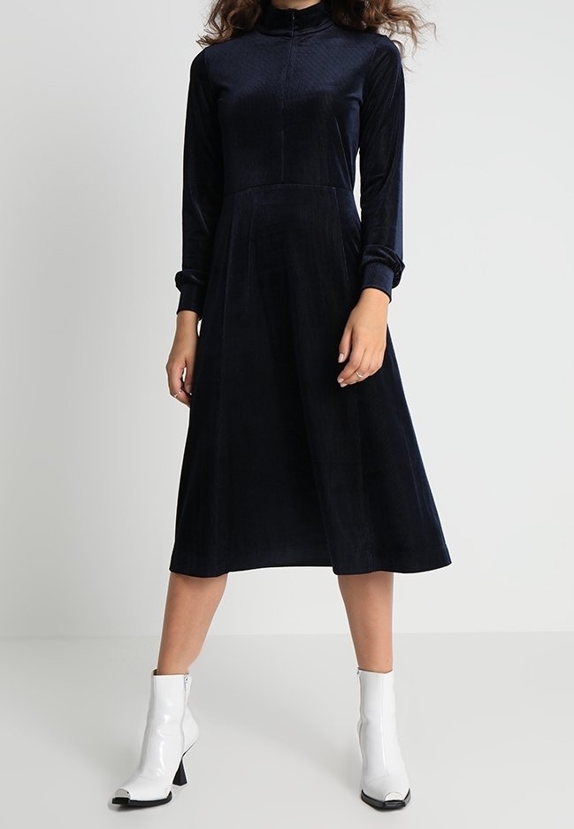 Femme vêtue d'une robe midi noire à manches longues avec un col montant, associée à des bottines blanches, debout devant un fond clair et uni.