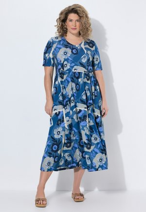 Mujer con cabello rizado que lleva un vestido midi azul con flores y mangas cortas, de pie y usando sandalias de cuña marrones contra un fondo blanco.