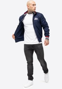 Mørkeblå bomberjakke med opretstående krave, røde og hvide striber, lynlås lukning, båret over en hvid t-shirt og sorte jeans.