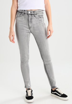 Personne portant un jean skinny gris clair, un haut court blanc et des baskets noires avec des semelles et des lacets blancs, debout sur un fond blanc.
