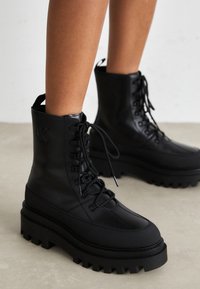 Bottes de combat en cuir noir avec une semelle épaisse, un fermoir à lacets à l'avant et des accents texturés ; présente un logo en relief et une conception robuste.