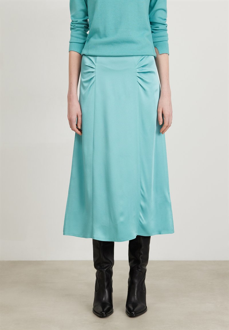 Une jupe satin aqua blue mi-longue avec des détails froncés sur les côtés, associée à des bottes noires à talons jusqu'aux genoux. Texture lisse, silhouette en A.