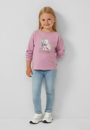 Maglietta a maniche lunghe rosa chiaro con un grafico di un gatto al centro e del testo, abbinata a jeans azzurri e sneakers bianche.