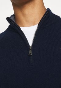 Granatowy dzianinowy sweter z suwakiem sięgającym do ćwiartki długości i kołnierzem typu stójka. Charakteryzuje się prążkowaną fakturą oraz białą wewnętrzną warstwą widoczną przy kołnierzu.