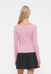 Rosa langermet topp med bred U-hals og ribbet tekstur, kombinert med en plissert svart skjørt. Bakfra vises tilpassede designdetaljer.