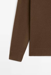 Manica e parte bassa di un maglione lavorato a maglia marrone, con polsini e orlo a coste, su sfondo bianco.