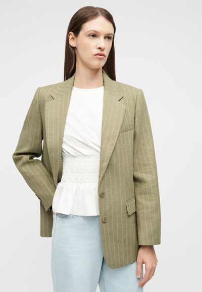 Femme portant un blazer beige à rayures fines sur une blouse blanche froncée et un jean bleu clair, debout devant un fond clair uni.