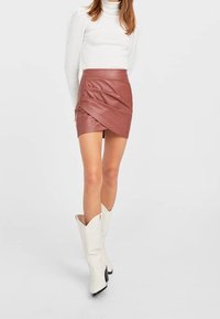 Mini-jupe marron en simili cuir avec un design drapé, associée à un col roulé blanc moulant et des bottes blanches montantes jusqu'aux genoux.