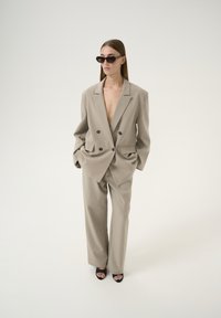 Beige, doppelreihiger Blazer mit großen schwarzen Knöpfen, kombiniert mit weit geschnittenen Hosen. Die Accessoires umfassen schmale schwarze Sonnenbrillen und Sandalen mit Absatz.