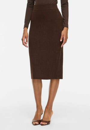 Mujer que lleva una falda lápiz ajustada de color marrón oscuro hasta la rodilla con cintura alta, combinada con tacones marrones abiertos y una blusa de manga larga.
