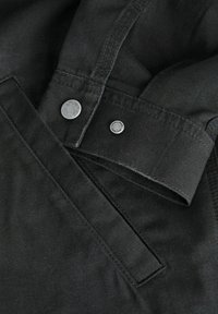 Gros plan d'une veste en tissu noir montrant un poignet boutonné et une poche inclinée avec des coutures visibles et des boutons-pression métalliques.